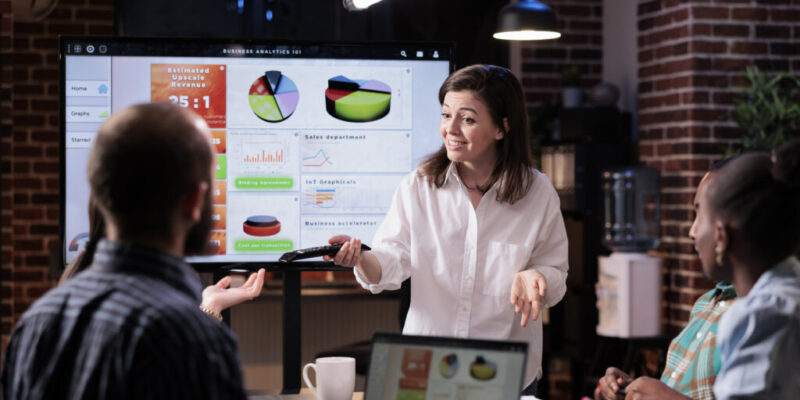 Smiling caucasian woman in startup office doing business presentation on big tv screen with charts